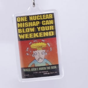 May include: A yellow and black keychain with a cartoon illustration of a person with a mushroom cloud over their head. The text reads "One nuclear mishap can blow your weekend.  Nukes aren't worth the risk.  1991 Jim Benton.  Southfield, Troy, MI."