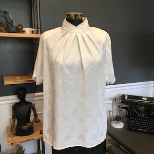 Puede incluir: Una blusa blanca de satén con cuello alto y mangas cortas. La blusa tiene un estampado de paisley.