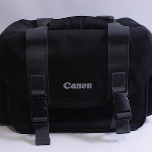 May include: Black Canon camera bag with adjustable straps and a zipped pocket. The bag has a silver Canon logo on the front.