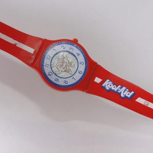 May include: A bright red wristwatch featuring a white stripe along the band and the Kool-Aid logo printed in blue and white. The round watch face has a blue border with black numbers from 1 to 12 and a central graphic of the Kool-Aid character.
