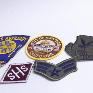 May include: A collection of embroidered patches with various designs and text. Includes a blue and yellow "Ladies Auxiliary" patch, a circular "City of Hanford" patch, and a military insignia.