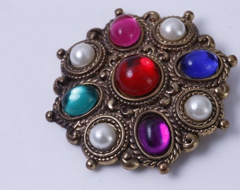 Könnte beinhalten: Eine goldfarbene Brosche mit einem Cluster aus bunten Edelsteinen und Perlenimitationen. Die Brosche zeigt einen roten, rosa, blauen, grünen und violetten Edelstein in einem kreisförmigen Design.