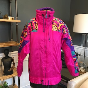 May include: A pink ski jacket with a purple and blue geometric pattern. The jacket has a high collar and a zipper closure.