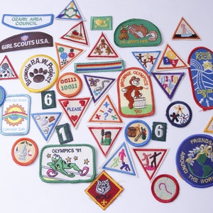 May include: A collection of colorful Girl Scout badges featuring various designs, including a paw print, a frog, a wolf, and a globe. The badges are made of fabric and have embroidered details. The text on the badges includes "Girl Scouts USA", "Brownie Olympics", "Going Places", and "Friendships Around the World".