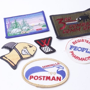 Puede incluir: Una colección de parches bordados, que incluyen una escena de un campamento, un parche militar con el texto "3rd Combat Comm GP", un parche con el texto "Registered Peoples Pharmacist", un parche con el texto "Skirmish U.S.A. 1999 Postman", un parche de dibujos animados de un pez amarillo con contorno negro, y un parche con una "W" blanca y una bandera a cuadros.