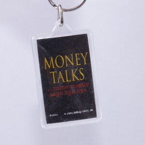 Może przedstawiać: Czarno-żółty brelok do kluczy z tekstem "MONEY TALKS...mine mostly says bye-bye." i informacją o prawach autorskich "© 1991, Self-Up, TROY, MI".