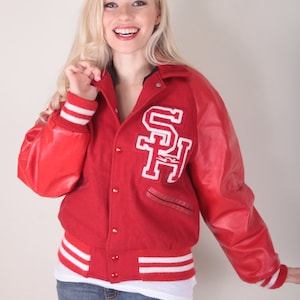 Könnte beinhalten: Eine rote und weiße Collegejacke mit Lederärmeln. Die Jacke hat einen weißen Filzbuchstaben-Patch auf der linken Seite, auf dem "S.F.H." steht.
