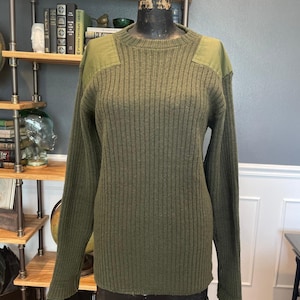 Puede incluir: Jersey de punto acanalado verde oliva con parches reforzados en los hombros. El jersey de manga larga se muestra en un maniquí. El jersey tiene cuello redondo y un diseño clásico de inspiración militar.
