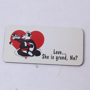 May include: A rectangular white tag with rounded corners featuring a graphic of two cartoon skunks embracing in front of a red heart. The text "Love... She is grand, No?" is printed in black on the right side. A small circular hole is on the left.