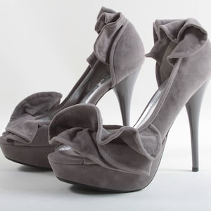 Puede incluir: Un par de zapatos de tacón alto de gamuza gris con un detalle de volantes alrededor del tobillo y la punta.