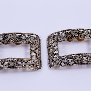 Peut inclure: Deux boucles métalliques ornées d'un motif de filigrane. Les boucles sont de couleur dorée et ont une forme rectangulaire avec un dessus et un bas arrondis.