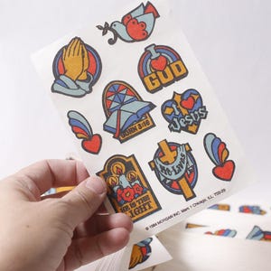 Puede incluir: Hoja de pegatinas religiosas vintage con varios símbolos como manos rezando, palomas, corazones y cruces. Las pegatinas tienen una paleta de colores retro de rojo, azul, amarillo y naranja. Algunas pegatinas incluyen texto como "GOD", "Jesus" y "He is the Light".