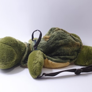 May include: A plush green turtle backpack with a brown strap. The turtle has a textured shell and a soft, fuzzy body.