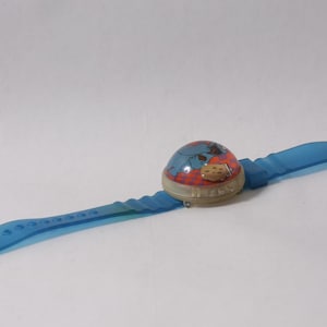 May include: A wristwatch featuring a translucent blue plastic strap and a domed, circular face. The watch face displays a graphic of a blue character with red accents and a small, light-colored circular detail, secured by a silver-toned metal buckle.