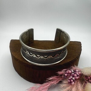 May include: A silver-toned cuff bracelet with intricate geometric patterns. The wide band features a textured design with a series of wavy lines and a woven-like pattern. The bracelet is displayed on a small, dark wooden block.