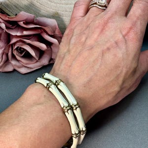 May include: Two cream-colored bangle bracelets featuring a segmented bamboo-like design with metallic gold-toned accents at each joint. The bracelets have a smooth, glossy finish and are worn together on the wrist, creating a stacked appearance.
