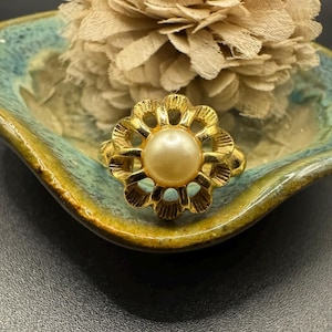 May include: A gold-toned ring with a central pearl-like bead. The ring has a floral design around the bead. The ring is set on a small, turquoise and gold-colored dish, with a beige flower in the background. Jewelry.