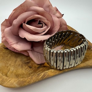 May include: Silver-toned metal stretch bracelet with a bamboo-style design. The bracelet is displayed on a wooden tray next to a large, dusty rose-colored artificial rose. The background is white.