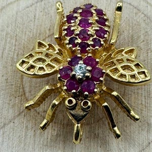 Puede incluir: Un colgante de insecto dorado con detalles intrincados. El cuerpo del insecto está adornado con numerosas piedras preciosas pequeñas, redondas y de color rojo intenso. Las alas tienen un diseño calado delicado. Una pequeña gema azul claro está en el centro del pecho del insecto.