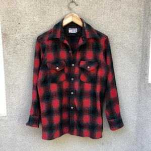 Könnte beinhalten: Ein rot-schwarz kariertes Flanellhemd mit zwei Brusttaschen und Knopfverschlüssen. Das Hemd hängt an einem Holzbügel vor neutralem Hintergrund. Der Kragen ist schwarz.