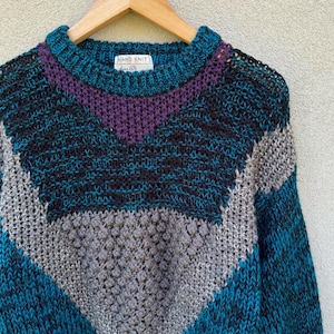 Puede incluir: Un suéter tejido a mano con un patrón geométrico en tonos turquesa, morado, gris y negro. El suéter tiene cuello redondo y está hecho con un hilo grueso. La etiqueta dice "Hand Knit".