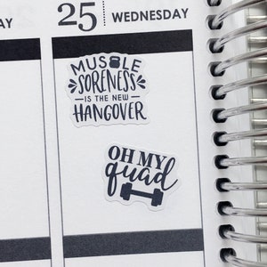 Op de afbeelding: Twee witte stickers met zwarte tekst en een haltergrafiek. De eerste sticker zegt "Muscle Soreness - Is the new - Hangover". De tweede sticker zegt "Oh My Quad" met een haltergrafiek.