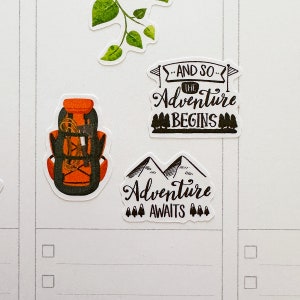 May include: A collection of adventure-themed stickers. One sticker depicts an orange and black backpack with rope detail. Other stickers include the phrases "And So The Adventure Begins" and "Adventure Awaits" with mountain and tree illustrations.