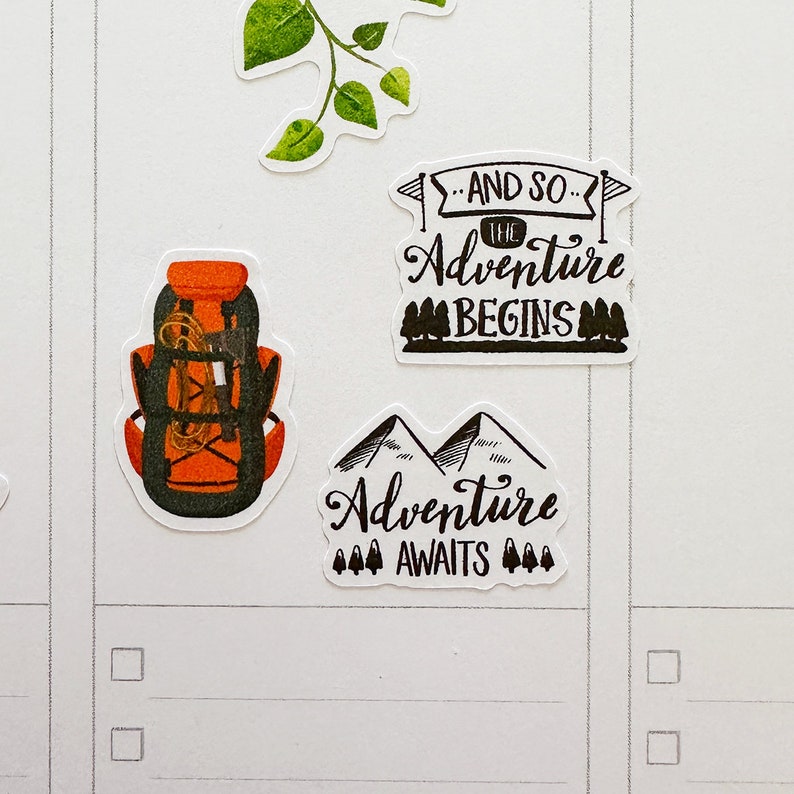 May include: A collection of adventure-themed stickers. One sticker depicts an orange and black backpack with rope detail. Other stickers include the phrases "And So The Adventure Begins" and "Adventure Awaits" with mountain and tree illustrations.