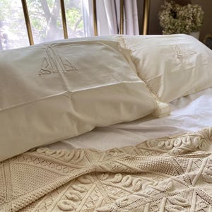 May include: Two cream-colored pillowcases featuring an embroidered monogram design and lace trim edges, paired with a cream-colored knit throw blanket displaying a textured geometric pattern with leaf-like motifs across the surface.