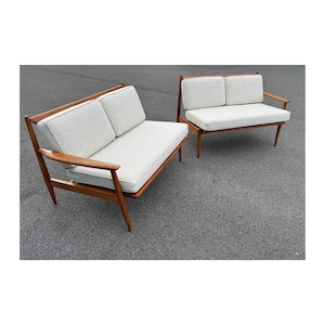May include: Two mid-century modern style sofas with light-colored cushions and wooden frames. The sofas have a two-seat design and a single-seat design. The wooden frames have angled legs and armrests. The cushions are a light, neutral color.