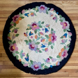 May include: A round rug with a cream-colored center and a dark blue border. The rug features a floral design with pink, purple, orange, and blue flowers. The rug is placed on a wooden floor.