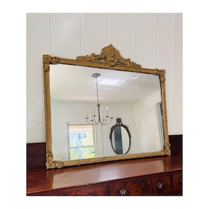May include: A large, rectangular antique mirror with a gold-colored ornate frame. The mirror is resting on a dark wood dresser. The frame has detailed carvings at the top and along the edges, adding to its vintage appeal.