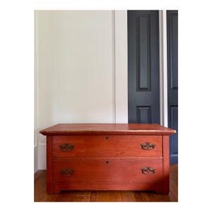 May include: A two-drawer antique chest of drawers with a reddish-orange finish. The chest has ornate brass handles and keyholes. It is set against a white wall and a dark blue door.