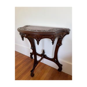 May include: A dark brown wooden console table featuring a semi-circular top with intricate carved detailing along the edges. The table is supported by three slender, curved legs that meet at a central point, accented with small decorative finials.