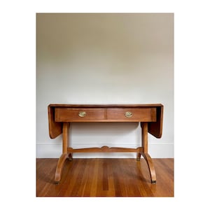 May include: A light brown wooden drop-leaf table featuring two drawers with circular brass pulls. The table has curved legs connected by a central arched stretcher and two hinged side leaves that are currently in the folded downward position.