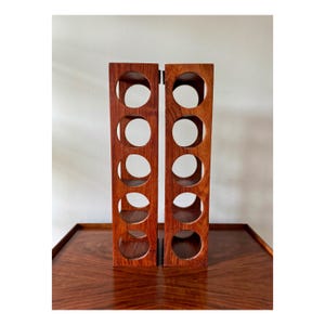 May include: Two wooden wine racks, each with five circular openings, are displayed side-by-side. The racks are a rich, reddish-brown color and appear to be made of solid wood. The design is minimalist and modern, suitable for storing wine bottles.