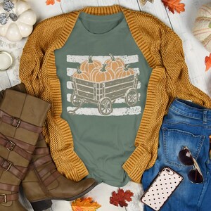 Pode incluir: Uma t-shirt verde sálvia com um gráfico de uma carroça de madeira cheia de abóboras, sob um cardigã de malha amarelo mostarda. O conjunto inclui calças de ganga azuis, botas castanhas até ao joelho com fivelas e uma capa de telemóvel às pintas.