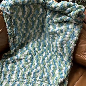Könnte beinhalten: Eine weiche, dicke Strickdecke in den Farben Weiß, Türkis und Grün. Die Decke ist aus einem flauschigen Garn gefertigt und hat ein strukturiertes, gewebtes Muster.