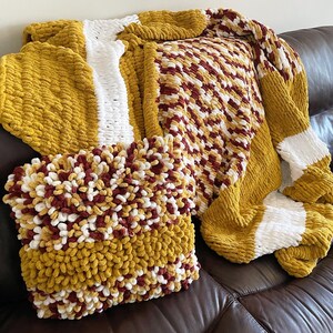 Könnte beinhalten: Ein handgefertigtes Decken- und Kissenset in Gold-, Weiß- und Burgundertönen. Die Decke hat ein gestreiftes Design und eine strukturierte, grobe Strickart. Das Kissen hat ein ähnliches strukturiertes Design. Die Artikel sind auf einem braunen Ledersofa ausgestellt.
