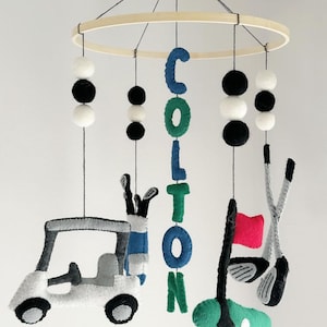 Può includere: Un mobile per neonati in feltro a tema golf. Presenta un golf cart, mazze da golf, una bandiera da golf e il nome "Colton" scritto in lettere di feltro. Il mobile è sospeso da un anello di legno e ha pompon neri e bianchi.