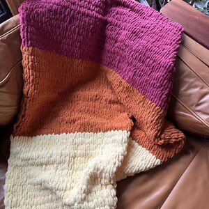 Könnte beinhalten: Eine dicke Strickdecke mit drei horizontalen Streifen in den Farben Burgunder, Orange und Creme.