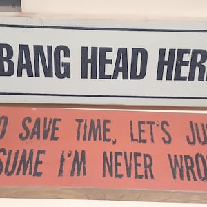 May include: Two rectangular wooden signs. The top sign is white with a black border and the words "BANG HEAD HERE" in black. The bottom sign is orange with black text that reads "TO SAVE TIME, LET'S JUST ASSUME I'M NEVER WRONG."