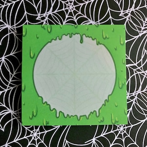 May include: Green sticky note with a white circle in the center. The circle has a spiderweb design. The green background is dripping with slime.