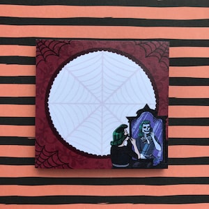 May include: A red and white notepad with a spooky illustration of a woman applying makeup while looking in a mirror. The mirror reflects a skeleton with a similar pose. The background is a dark red with a spiderweb design.