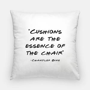 May include: White pillow with black text that reads "CUSHIONS ARE THE ESSENCE OF THE CHAIR" - CHANDLER BING