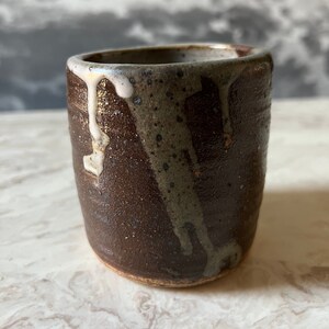 May include: A brown ceramic mug with a white glaze dripping down the side. The mug has a rustic, handmade look.