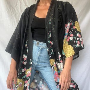 May include: A black kimono-style robe featuring a floral pattern with pink and white blossoms, gold geometric accents, and illustrated figures. The garment has wide sleeves, an open front, and a dark base color with subtle tonal script details.
