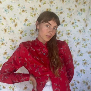 Puede incluir: Una camisa roja de manga larga con cuello mandarín y bordado de hojas. La camisa está abierta en la cintura y se usa con pantalones blancos. Se ve un pequeño collar con colgante azul.