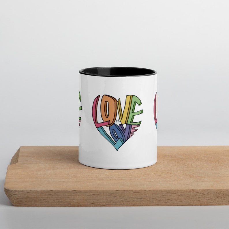 May include: White ceramic coffee mug with a black rim. The mug features a colorful graphic of a heart made of the word "LOVE" with the words "is LOVE" inside the heart.