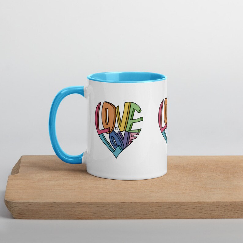 May include: White ceramic mug with a light blue rim and handle. The mug features a colorful graphic of the words "Love is Love" arranged in a heart shape.